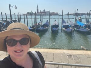 Life-Changing Travel Must-Haves for Women Over 50 1 Sue, wearing sunglasses and a sun hat, smiles in front of docked gondolas on a sunny day in Venice, with the city's historic buildings visible across the water.
