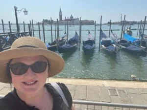 Sue, wearing sunglasses and a sun hat, smiles in front of docked gondolas on a sunny day in Venice, with the city's historic buildings visible across the water.
