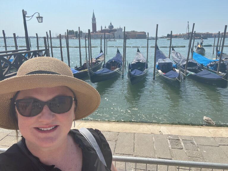 Life-Changing Travel Must-Haves for Women Over 50 8 Sue, wearing sunglasses and a sun hat, smiles in front of docked gondolas on a sunny day in Venice, with the city's historic buildings visible across the water.