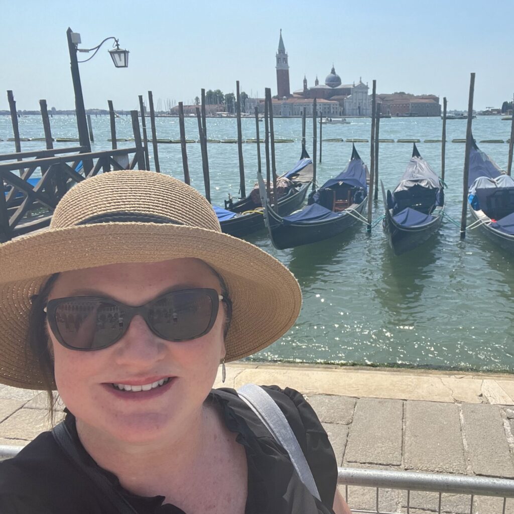 Sue, wearing sunglasses and a sun hat, smiles in front of docked gondolas on a sunny day in Venice, with the city's historic buildings visible across the water.