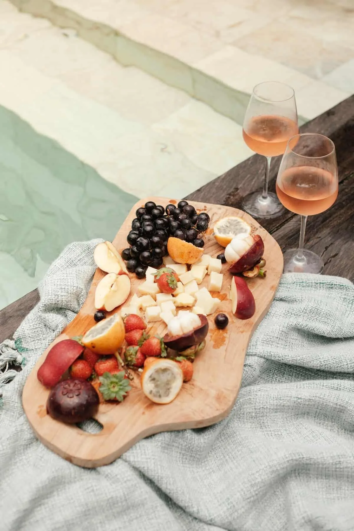 A wooden board with fruits and cheese by a pool, paired with two glasses of rosé wine.